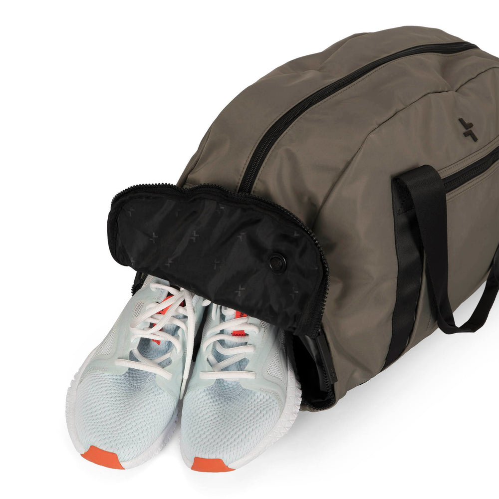 Angle view of a grey-green duffle bag called Sutton designed by Tracker on a white background, showcasing its top handle, front zipper pocket, main zipper compartment, and side seperate shoe pocket with shoes coming out of it.